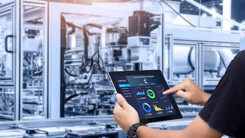 Um homem usa um tablet para monitorar a automação industrial em uma fábrica moderna, com máquinas e equipamentos ao fundo.